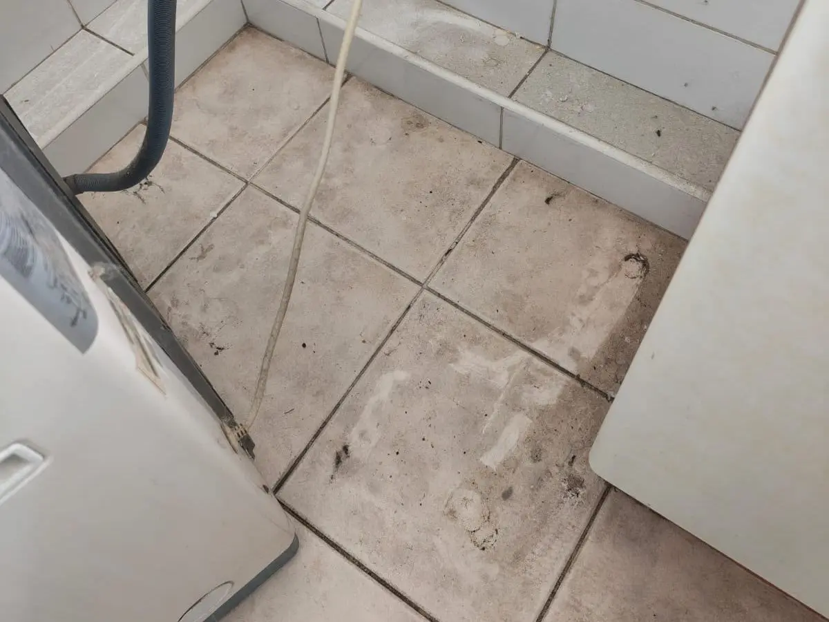 Piso de cerámica de una logia con manchas y suciedad acumulada bajo la lavadora, antes del aseo